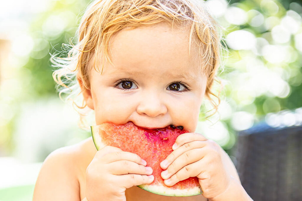 Nettes kaukasisches blondes Kind, das an einem heißen, sonnigen Sommertag Wassermelone isst. Baby Mädchen isst Früchte im Hinterhof. Bio-Wassermelone. Gesunde Gewohnheiten. Ausgeglichenes Leben. Langsames Leben. Sommerferien. Familienurlaub.Glückliche Kindheit. - Foto, Bild