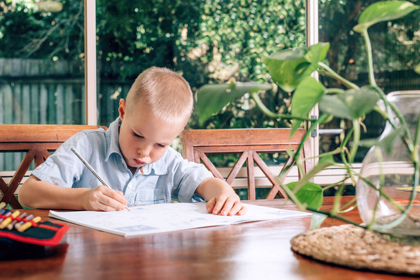 Pikkulapsi piirtää oppikirjaan kotona, poika pitelee kynää ja kirjoittaa. Hämärtynyt hirviökasvi etualalla
. - Valokuva, kuva