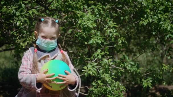 Portret młodej uroczej dziewczyny noszącej maskę ochronną z kulką koloru na tle drzew owocowych - Materiał filmowy, wideo