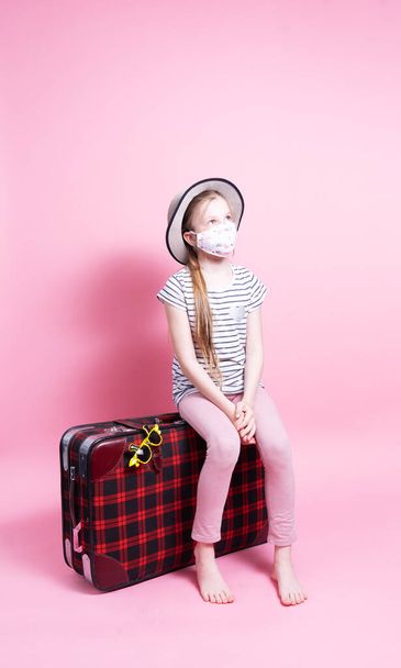Travel: A little girl in a straw hat and a medical mask is sitting on a big red suitcase and waiting for travel. - 写真・画像