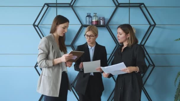 Tres mujeres se paran contra la pared en la oficina y verifican documentos para un proyecto empresarial
 - Metraje, vídeo