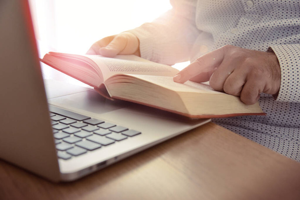 A man holds and reads a book in front of a laptop. Concept for education, business skill, personal and career development, online training and courses. - Photo, Image