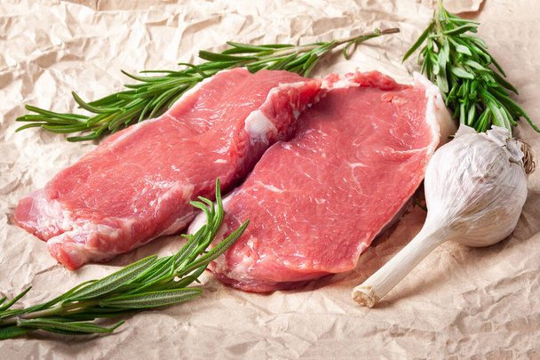 生肉とニンニク。紙の上に新鮮な生肉、ニンニク、ローズマリーの2枚のスライス。バーベキューのための肉の準備。健康的で自然食品. - 写真・画像