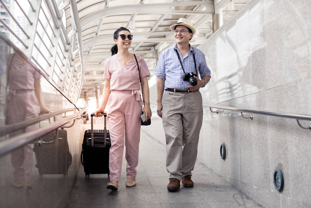 Idősebb ázsiai pár egy nővel, akik egy bőröndöt vonszolnak, és boldogan mosolyogva beszélgetnek a reptéren, hogy felkészüljenek az utazásra. A nagynénik és nagybácsik boldogsága együtt utaznak mosollyal. - Fotó, kép
