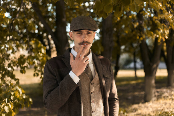 Retro retrato de 1920 de un gángster inglés con una gorra plana. Fuma un cigarrillo en la calle
.. - Foto, imagen