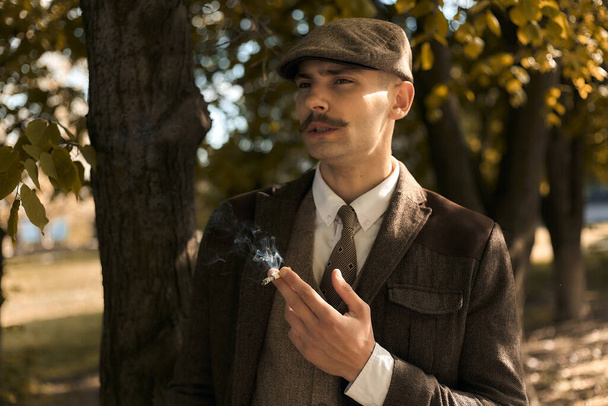 Portrait rétro des années 1920 d'un gangster anglais coiffé d'une casquette plate. fume une cigarette dans la rue
.. - Photo, image