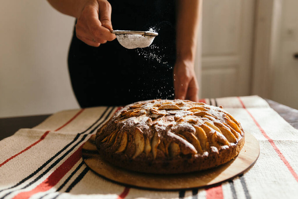 Woman hand sprinkles sugar powder on homemade apple cake. Cooking at home candid lifestyle concept. - Фото, изображение