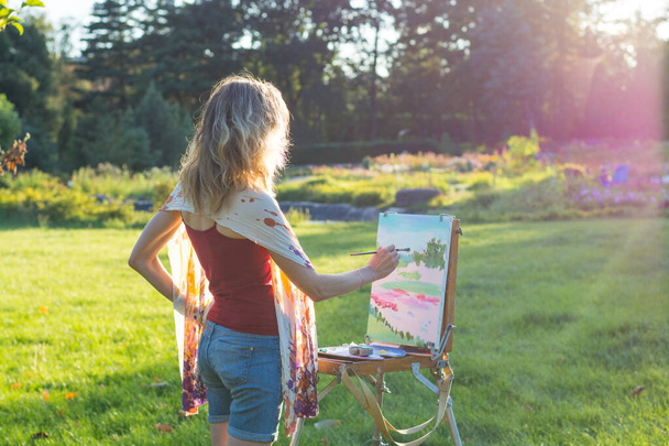 若い女性アーティストが筆を持ち、日没のイーゼルに絵を描く。画家は日没時に庭の油絵を描く - 写真・画像