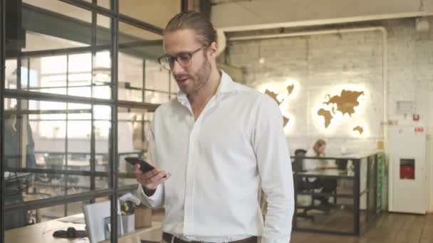 Skoncentrowany przystojny młody biznesmen w domu spacery w biurze za pomocą telefonu komórkowego - Materiał filmowy, wideo