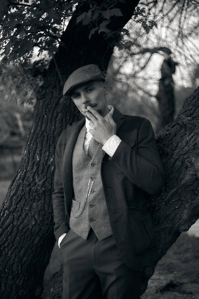 Retro-Porträt eines englischen Gangsters mit Schiebermütze. Raucht eine Zigarette auf der Straße.. - Foto, Bild
