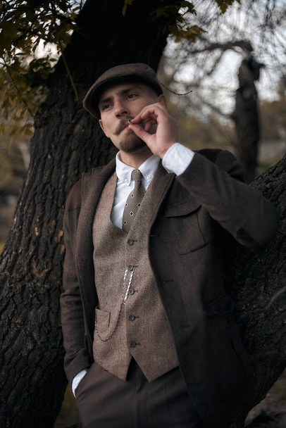 Ritratto retrò anni '20 di un gangster inglese con un cappuccio piatto. Fuma una sigaretta per strada
.. - Foto, immagini
