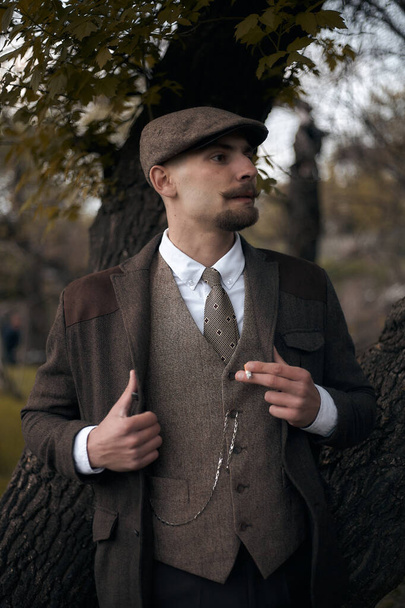 Retro 1920s portrait of an English gangster with a flat cap. Smokes a cigarette on the street.. - Фото, изображение