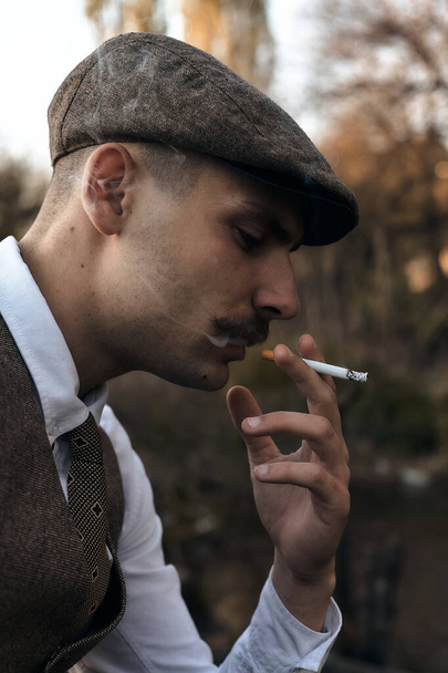 Portrait rétro des années 1920 d'un gangster anglais coiffé d'une casquette plate. fume une cigarette dans la rue
.. - Photo, image