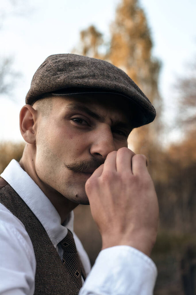 Retro 1920s portrait of an English gangster with a flat cap. Smokes a cigarette on the street.. - Фото, изображение