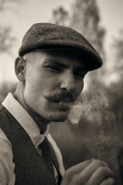 Retro-Porträt eines englischen Gangsters mit Schiebermütze. Raucht eine Zigarette auf der Straße.. - Foto, Bild