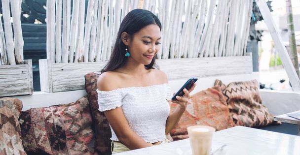 Beautiful young Asian female sitting with beverage in glass at table while smiling and using smartphone on terrace of cafe in Bali - Photo, image