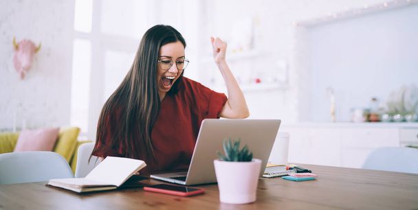 Elated excited successful female with long hair raising fist and screaming for celebrating victory watching on laptop at cozy apartment - Zdjęcie, obraz