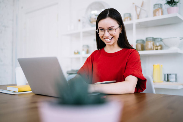 Felice ridendo donna casual in occhiali rotondi seduto di fronte al computer portatile e ridendo mentre messaggia sui social media durante la pausa  - Foto, immagini