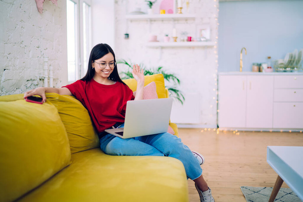 Attractive woman sitting on yellow couch and waving with hand while chatting via laptop having video chat sitting on couch at home  - Фото, изображение