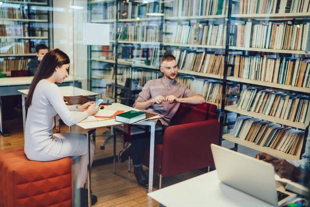 Spolupracovníci nosí formální obleky mluvit spolu a pomocí různých zařízení, zatímco sedí u stolů v knihovně v blízkosti knihoven - Fotografie, Obrázek