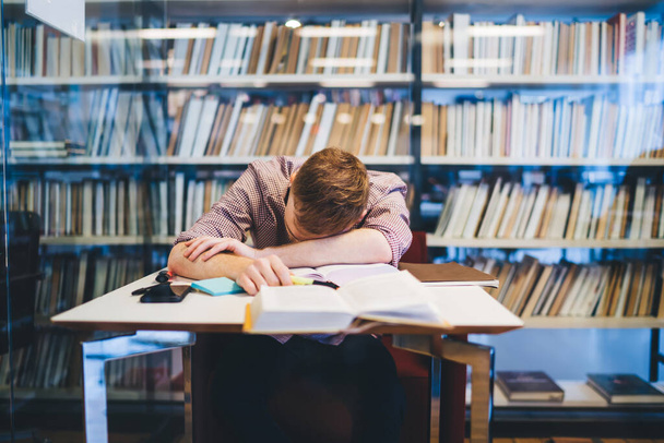 Exhausted male entrepreneur dressed in formal suit lying face down on table with books and notepads in library near bookshelves - Photo, Image