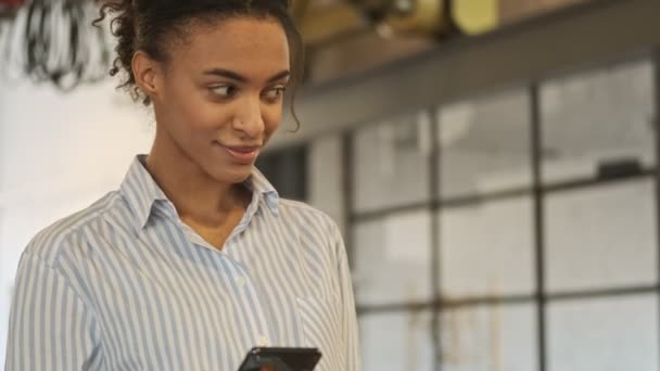 Θετική χαρούμενη νεαρή Αφρικανή επιχειρηματίας που μπαίνει στο γραφείο χρησιμοποιώντας κινητό τηλέφωνο - Πλάνα, βίντεο