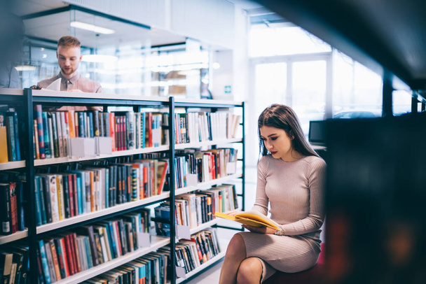 Young lady in trendy dress sitting near bookshelf and reading interesting book while doing homework in light library of university - Фото, изображение