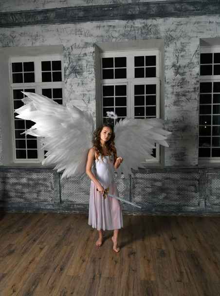κορίτσι στα λευκά με άσπρα φτερά αγγέλου με σπαθί ανταπόδοσης που ποζάρει σε γκρι φόντο - Φωτογραφία, εικόνα