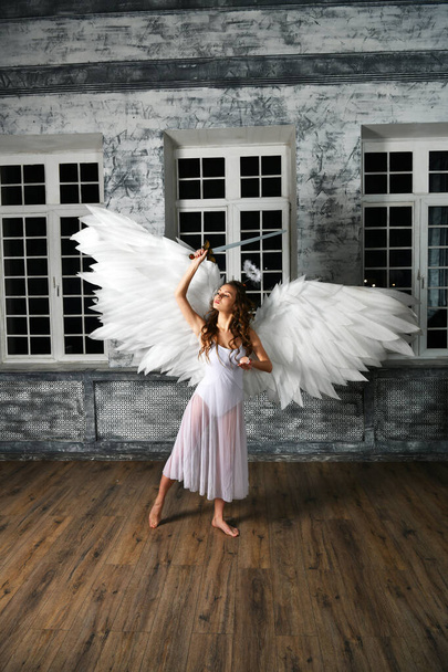 κορίτσι στα λευκά με άσπρα φτερά αγγέλου με σπαθί ανταπόδοσης που ποζάρει σε γκρι φόντο - Φωτογραφία, εικόνα