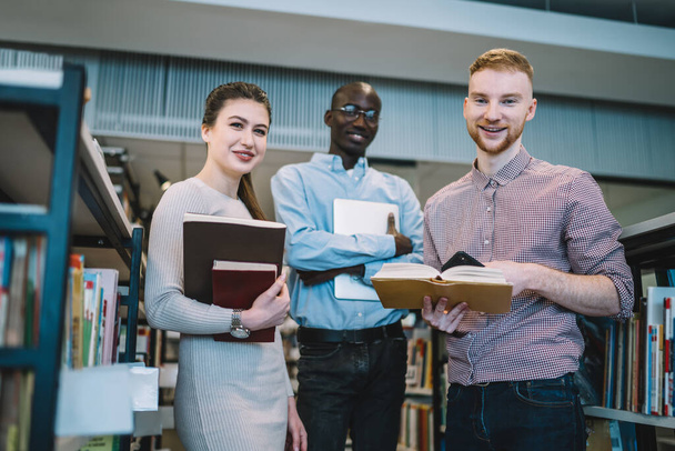 Confident smart multiethnic students in casual clothes holding notebooks and open books in bookstore and looking at camera with wide smile - Φωτογραφία, εικόνα