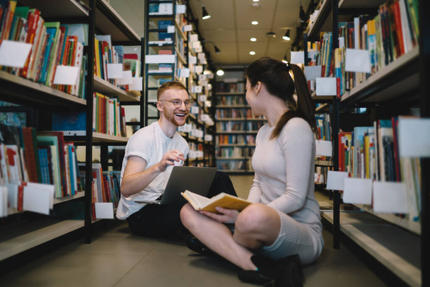 Delighted and happy redhead guy sitting on floor in library and using laptop while talking with lady reading interesting book - Фото, изображение