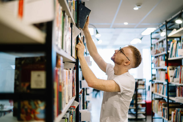 Side view of adult bearded man in white shirt and glasses concentrating while taking book from bookcase in light modern library - Foto, Bild