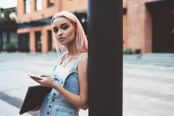 Serious young female with pink hair carrying sketchbook and looking at camera while leaning on pillar and browsing smartphone on street - Foto, Bild