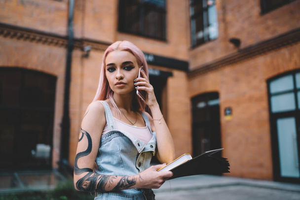 Stylish young lady with snake tattoo and long pink hair dressed in jeans dress speaking on mobile phone holding book in hand  - Zdjęcie, obraz