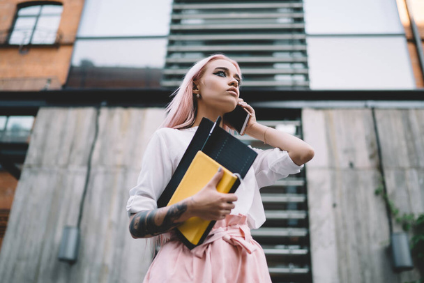 Low angle of young attractive tattooed female wearing trendy summer clothes having call on mobile phone while standing in street with notebooks and looking away - Foto, immagini