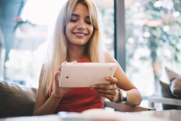 Glad young woman with blond hair cheerfully smiling and using modern tablet while sitting on sofa against window in cozy coffeehouse - Photo, Image