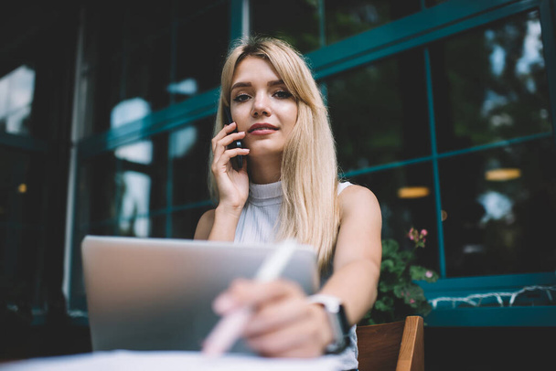 Блондинка усміхнена розумна леді в без рукавів сорочка розмовляє на мобільному телефоні, сидячи за столом з ноутбуком у вуличному кафе в місті
 - Фото, зображення