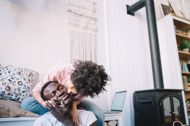Ethnic curly haired woman in striped shirt hugging from back and kissing on cheek happy African American man with eyes closed while sitting in living room with fireplace - Foto, Bild
