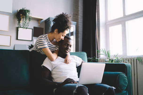 Glad African American female embracing ethnic man with laptop from back and smiling while standing behind comfortable couch and resting at home - Photo, Image