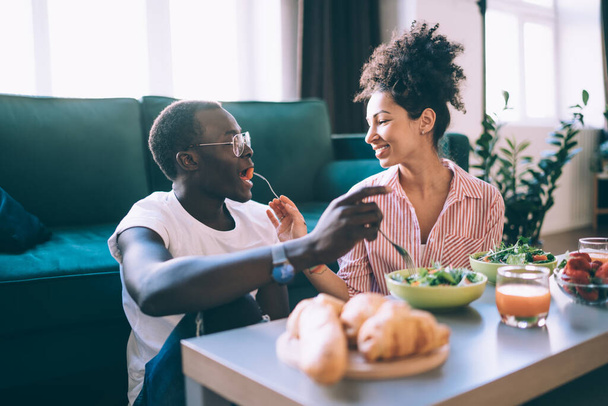 Smiling ethnic adult woman feeding pleased African American man in glasses while sitting on floor at table near sofa in light modern apartment on blurred background - Fotó, kép