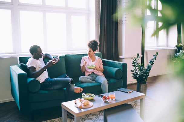 Black casual typical calm couple eating food on cozy big couch in apartment while husband browsing phone sitting opposite curly wife - Fotó, kép