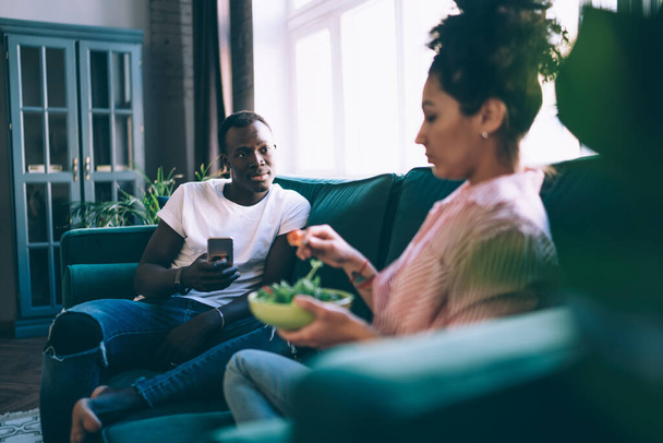 Serious young handsome African American man holding phone and looking at woman with bowl of salad while sitting together on cozy coach in apartment - Foto, afbeelding