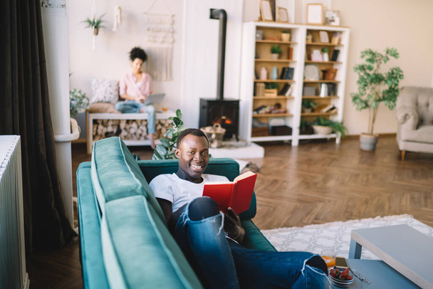 Multiracial stylish young couple comfortably placing on green sofa and bench with crossed legs for reading books in light spacious living room - Fotoğraf, Görsel