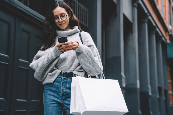 Pretty young woman in stylish sweeter and eyeglasses holding shopping bags browsing mobile while walking down street of New York city  - Foto, Bild