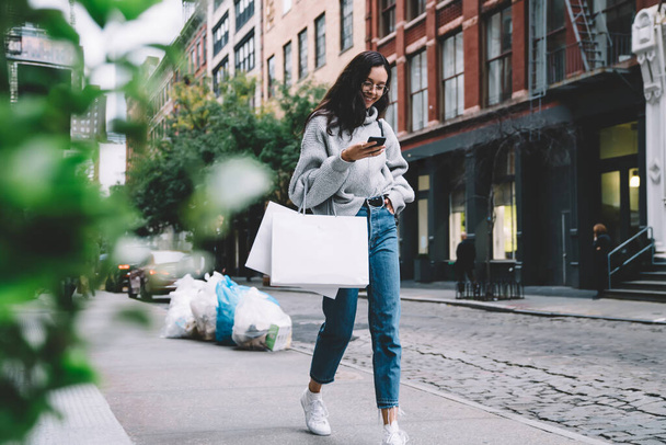 Beautiful modern woman in glasses carrying blank shopping bags wearing denim and sweater using smartphone while walking on city street - Valokuva, kuva