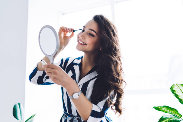 Charming tender dark haired woman smiling and applying mascara while holding and looking at hand mirror on background of window in apartment - Foto, afbeelding