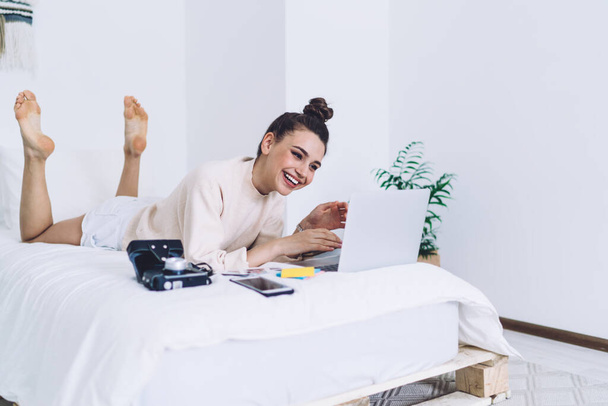 Joyful cheerful female social media marketing specialist spending time at home lying on stomach on bed with laptop and photo camera - Fotó, kép