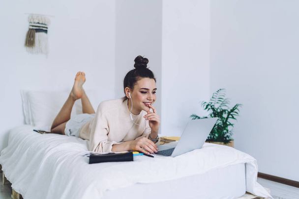 Thoughtful young woman with hair knot relaxing on bed with black purse browsing laptop touching lips and smiling in white room - Photo, image