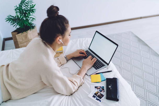 Side view of  casually dressed young woman with high bun typing new post for website on laptop with blank screen while lying on bed and enjoying good music - Фото, изображение
