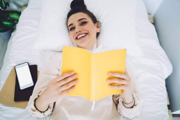 From above young charming laughing female with dark hair wrapped in bun relaxing at home and lying on bed with yellow book and smartphone - Photo, image